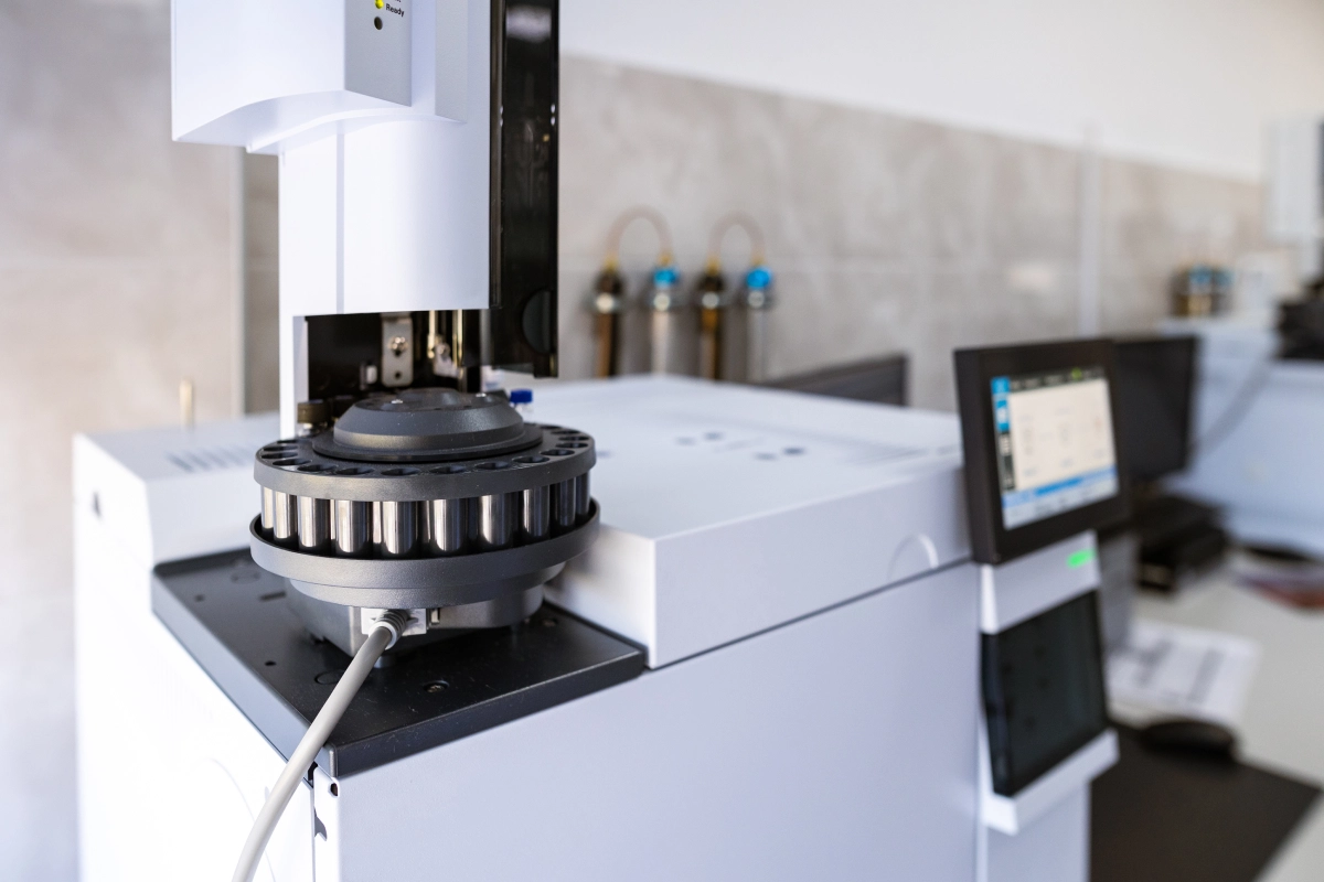 Laboratory equipment including a gas chromatography machine with a control panel and sample injector, situated in a lab with bottles on a shelf in the background.